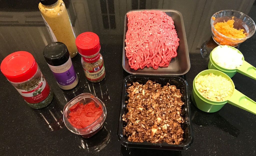 Jars of prepared mustard, parsley, garlic powder, and oregano; tomato paste; ground beef; finely chopped mushrooms; grated carrot in a custard cup; minced onion in a 1/2 cup measuring cup; and breadcrumbs in a 3/4 cup measuring cup